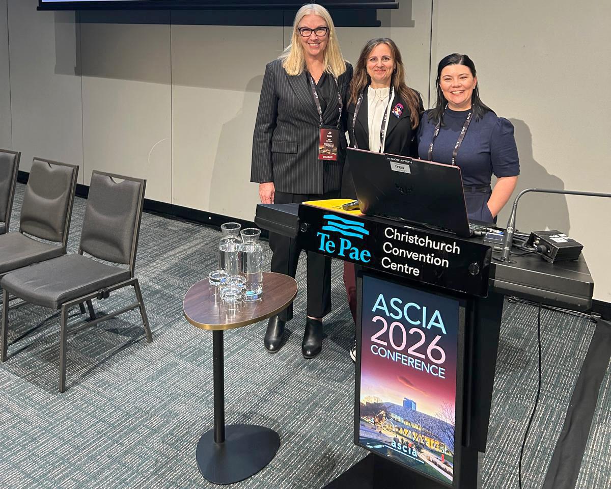 Three women standing at a podium with "ASCIA 2026 Conference" sign at the Te Pae Christchurch Convention Centre. Nearby are chairs and a table with water.
