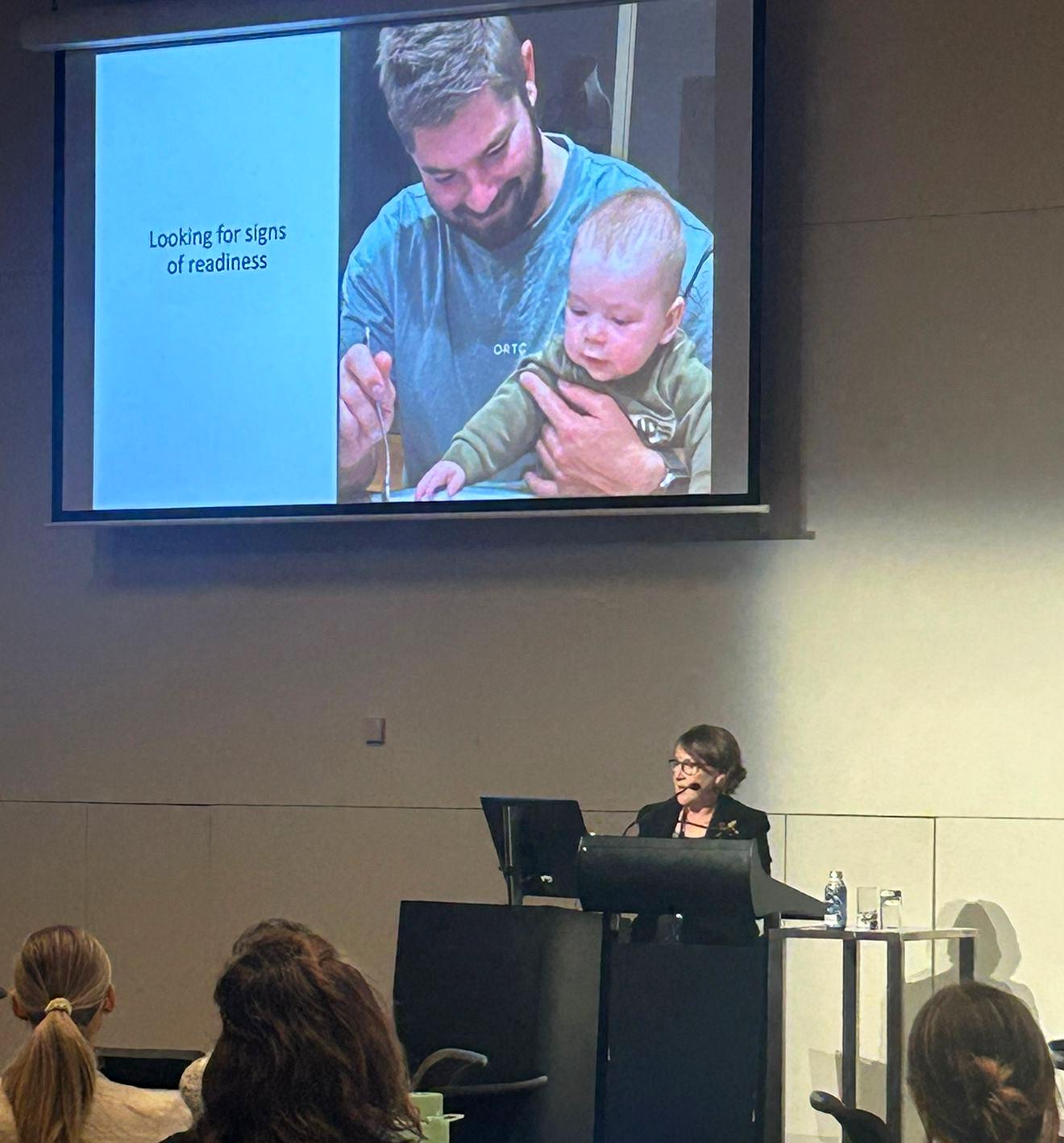 A speaker presents a slide showing a person with a baby, with the text "Looking for signs of readiness," to an audience in a lecture room.