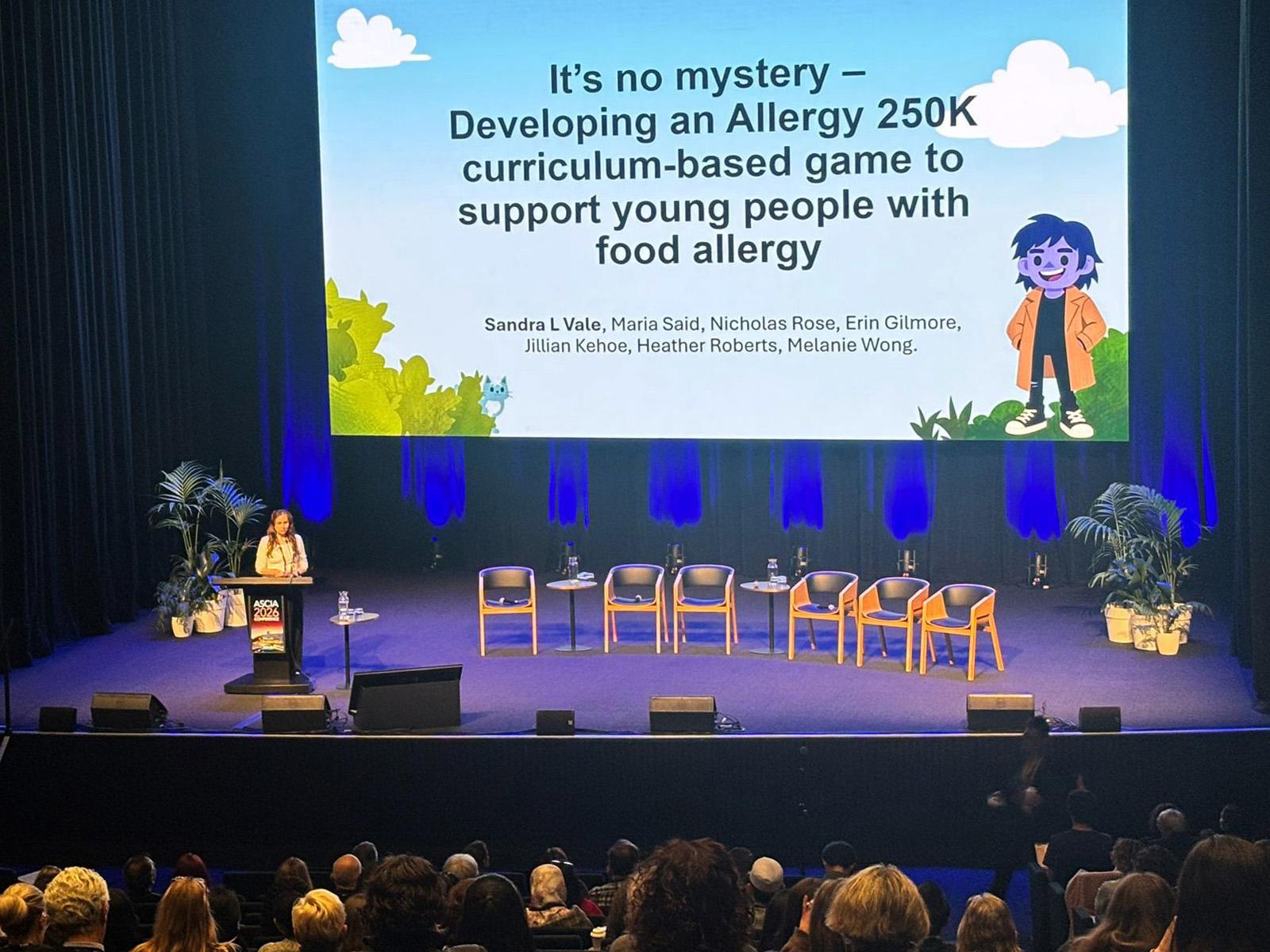 A woman speaks at a podium on stage with a large screen displaying a presentation about developing an allergy curriculum-based game. Audience visible.