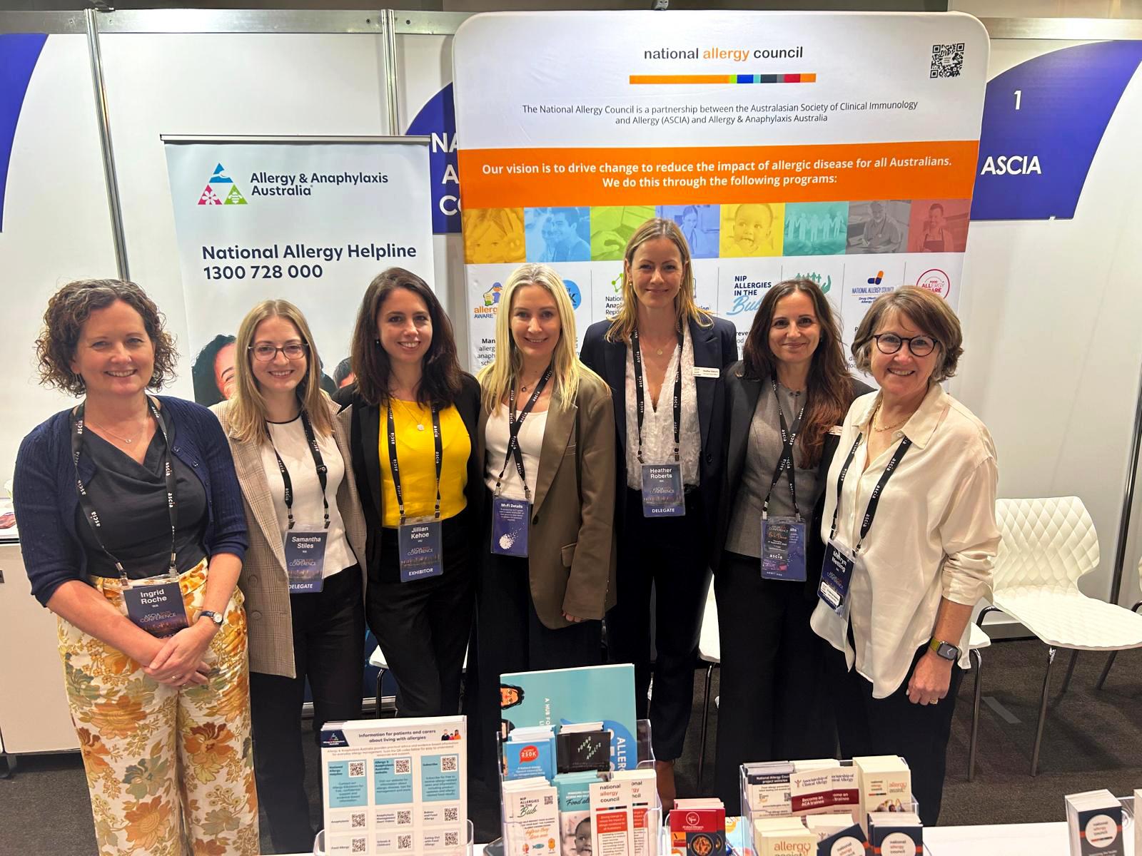 A group of seven people smiling and standing together at an allergy conference booth with informational displays and brochures.
