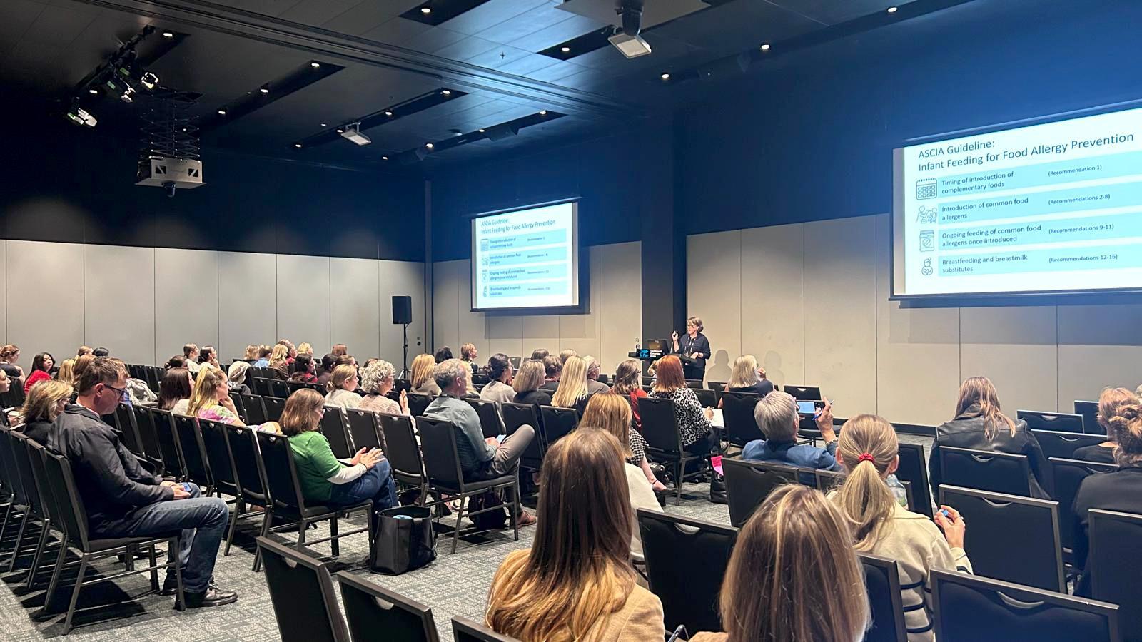 Audience seated in a conference room listens to a speaker presenting a slide titled "ASCIA Guideline: Infant Feeding for Food Allergy Prevention."