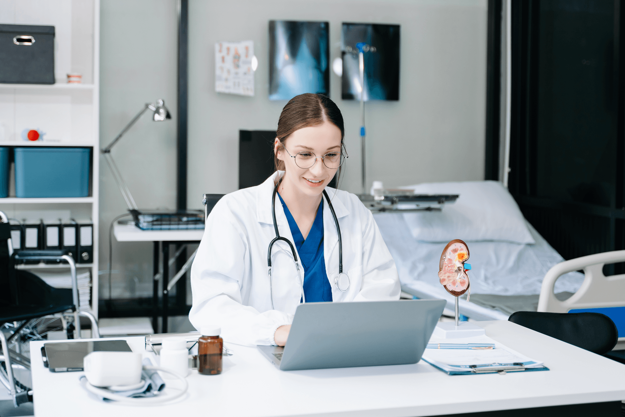 A doctor in a white coat and stethoscope works on a laptop at a desk in a medical office, with a kidney model and medical equipment nearby.