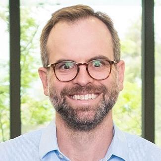 A smiling man with glasses, beard, and short hair, wearing a light blue shirt, stands in front of a window with greenery in the background.
