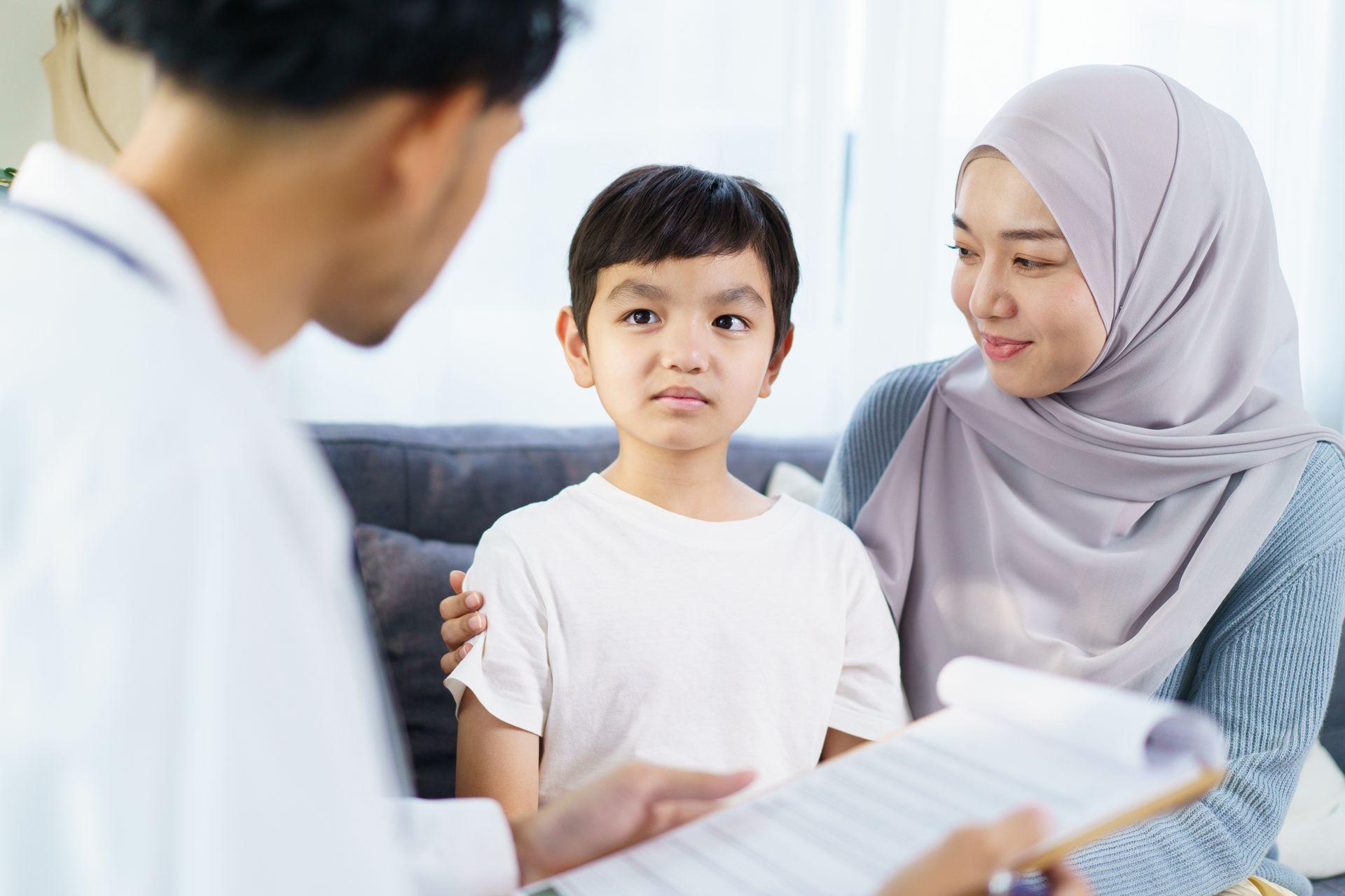 A child sits between a mother in a hijab and a doctor holding a clipboard, in a medical consultation setting.