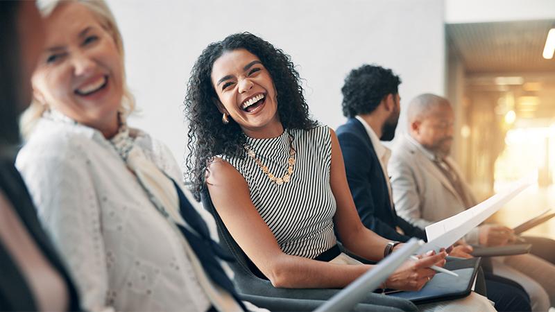 A group of people in business attire sitting and laughing together in a bright, modern office setting.