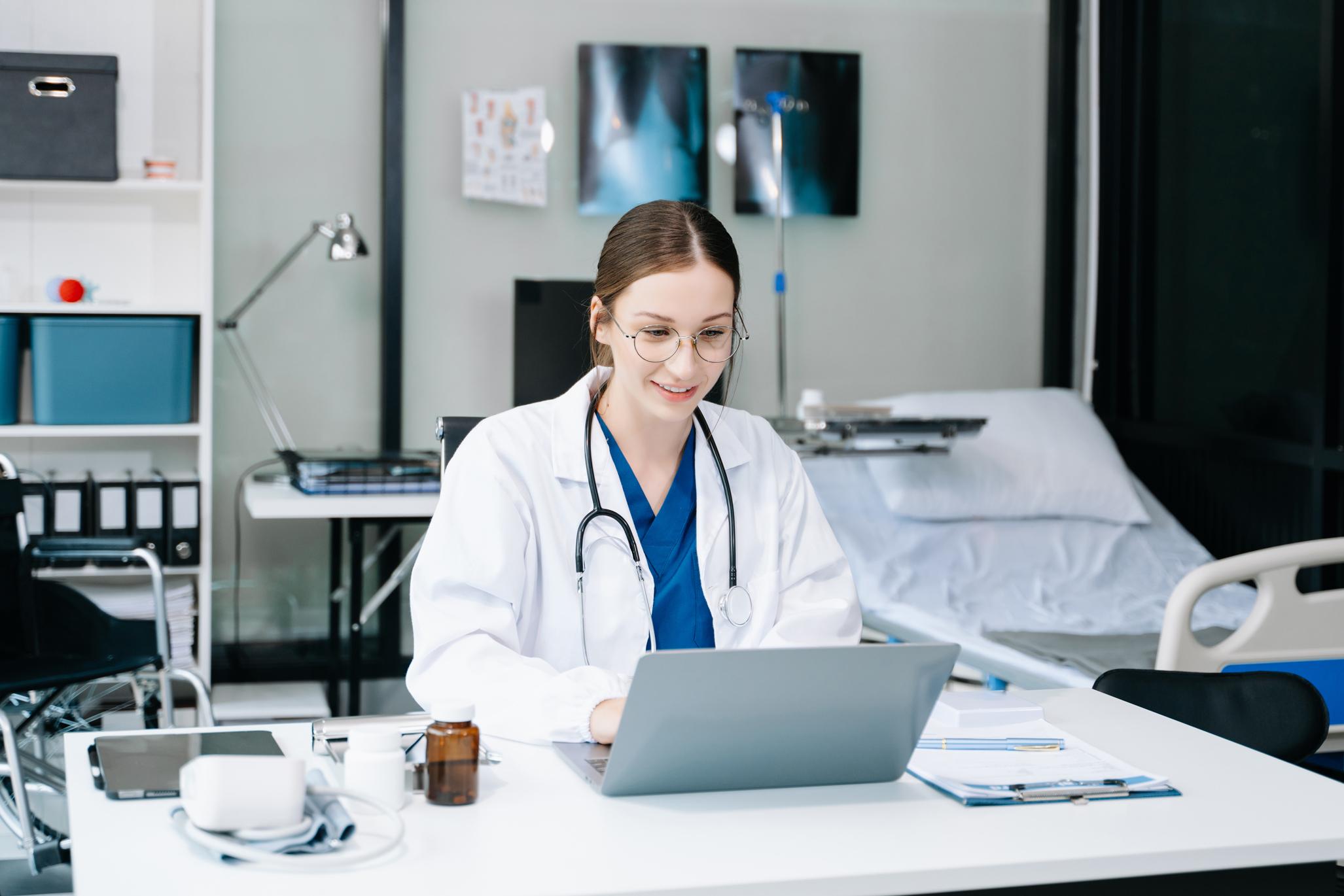 A doctor in a white coat and stethoscope works on a laptop at a desk in a modern medical office with a hospital bed in the background.