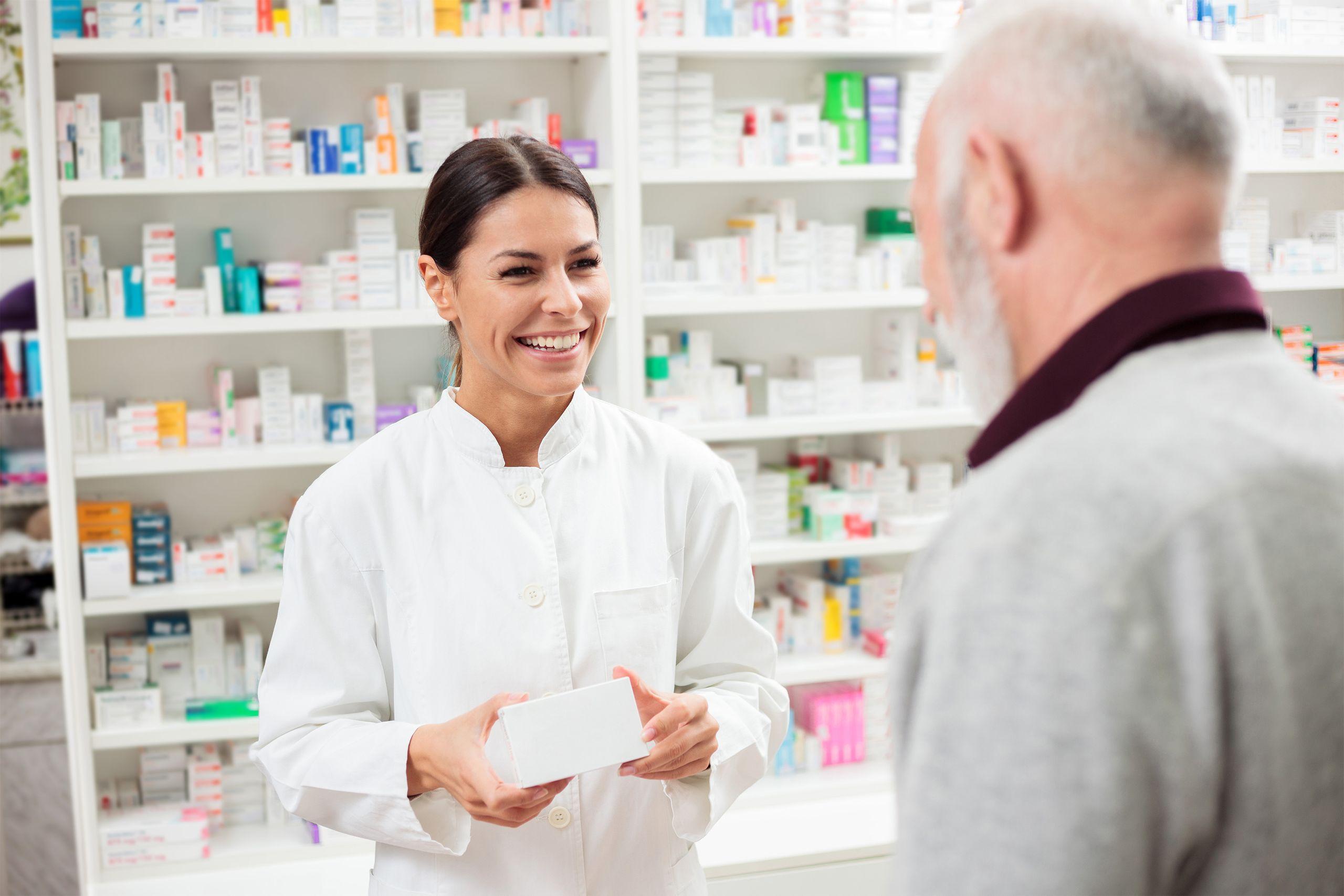 Pharmacist in a white coat smiling and handing a medication box to an older man in a pharmacy with shelves of medicine in the background.