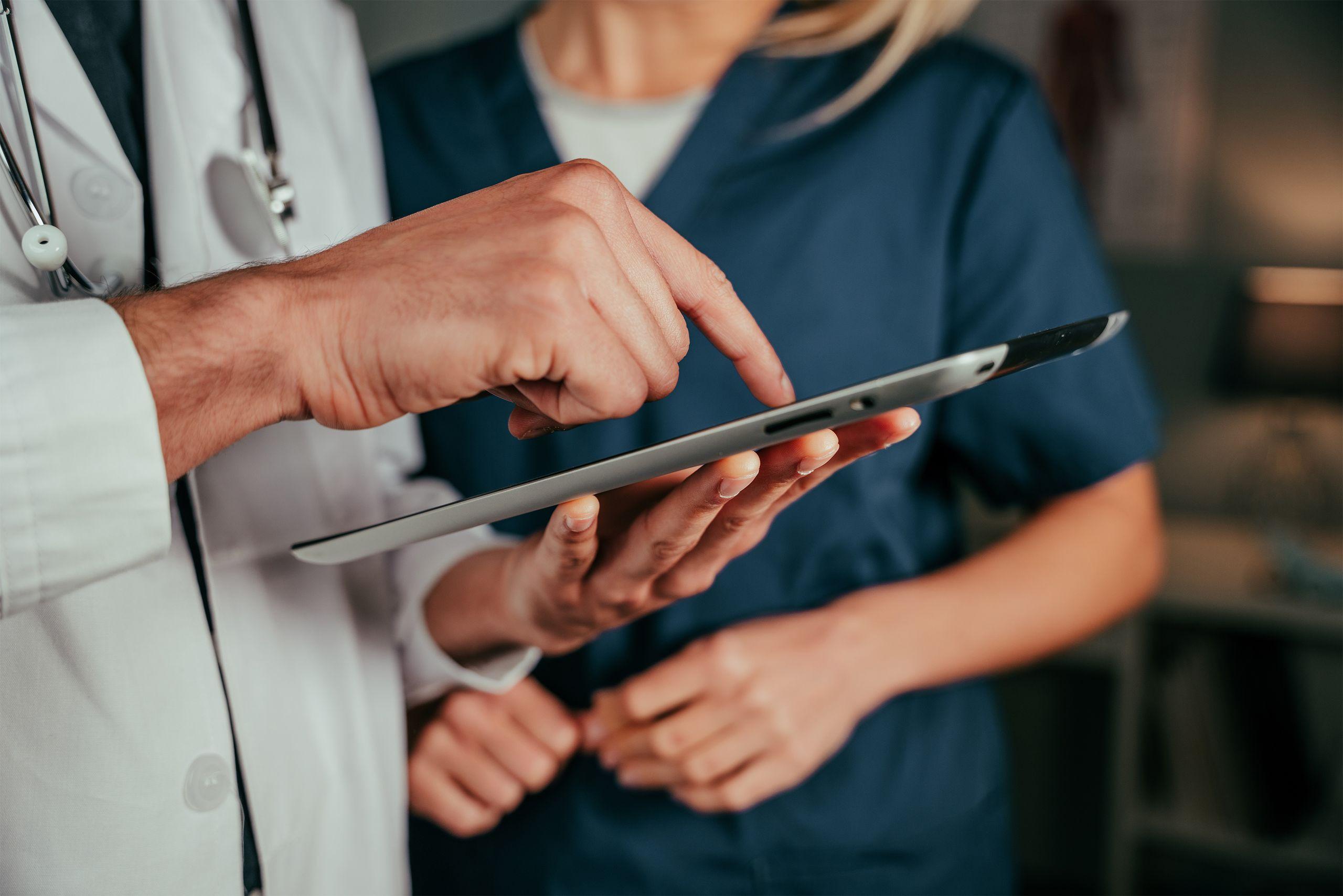 A doctor and nurse discuss information on a tablet, focusing on the screen. Both are in medical attire in a clinical setting.