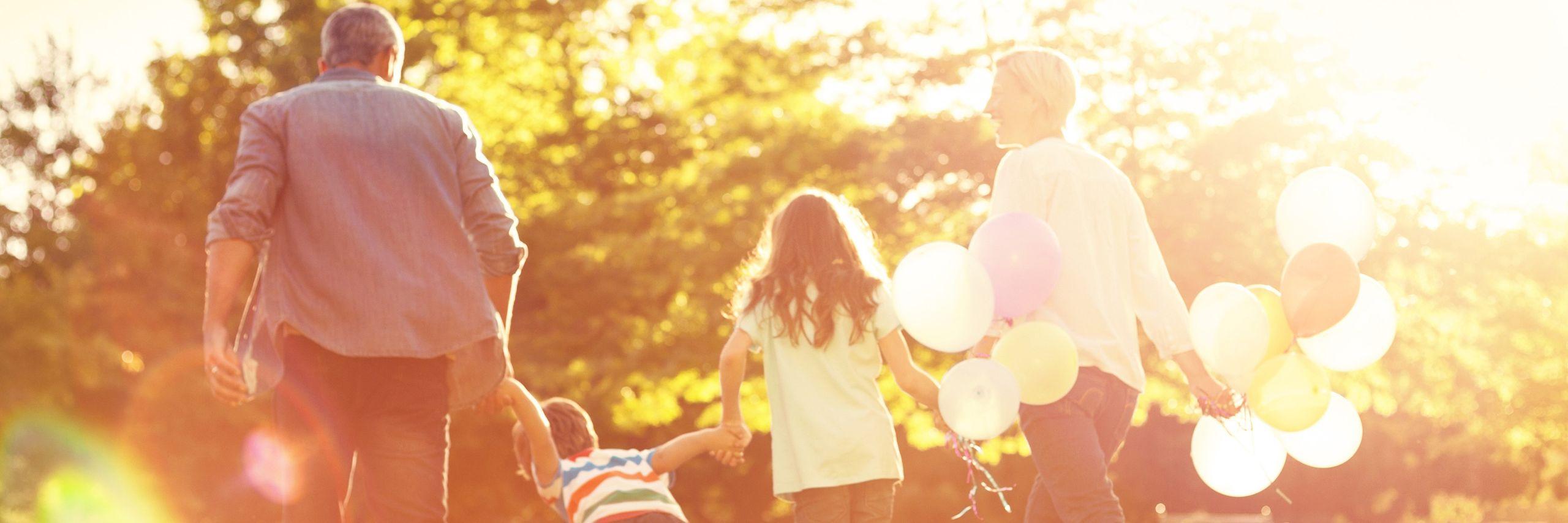 A family walks hand in hand outdoors, with a woman holding colorful balloons, bathed in warm sunlight.