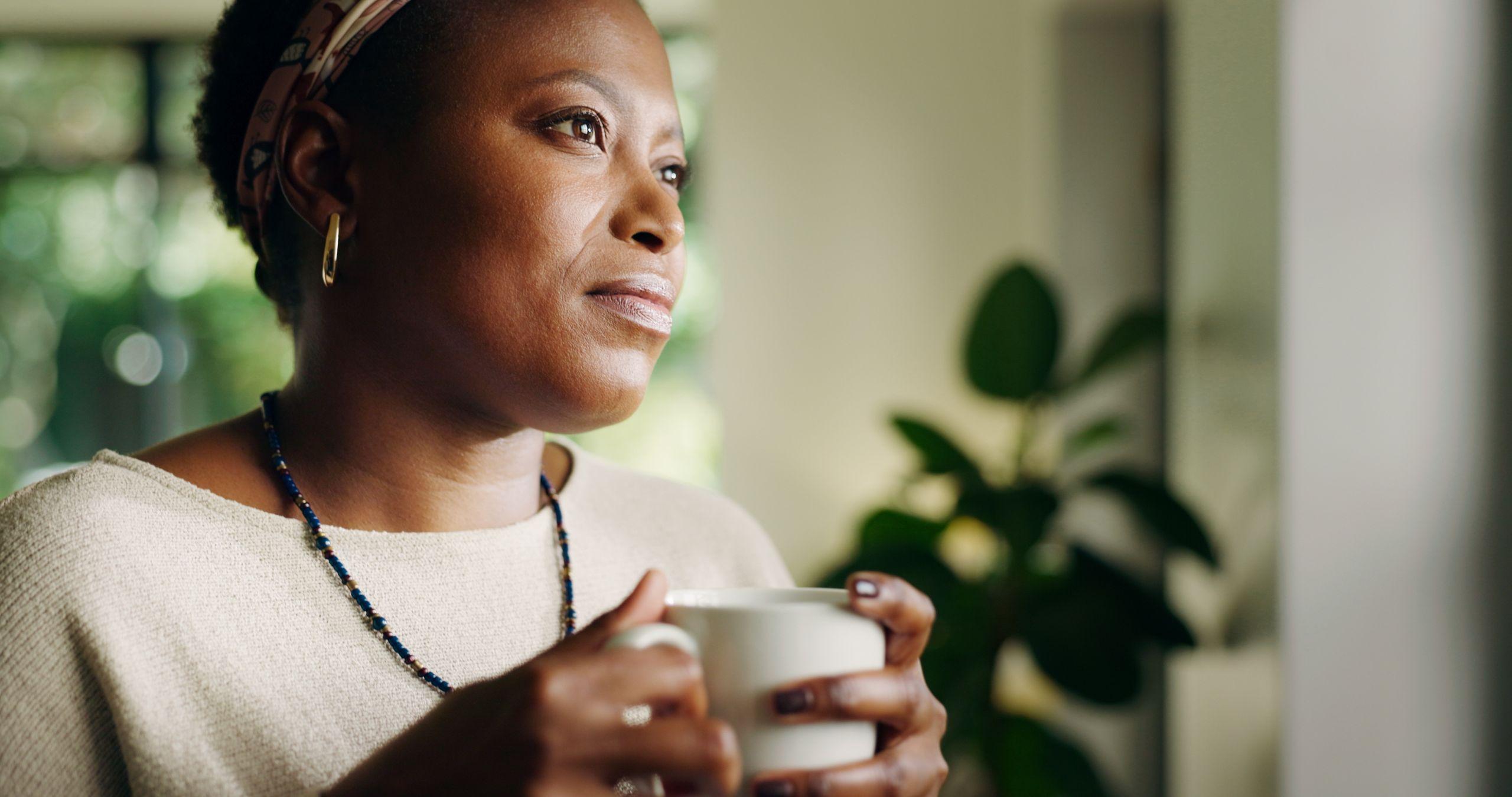 Woman with a thoughtful expression, holding a white mug, wearing a beige sweater and necklace, in a softly lit room with a plant in the background.