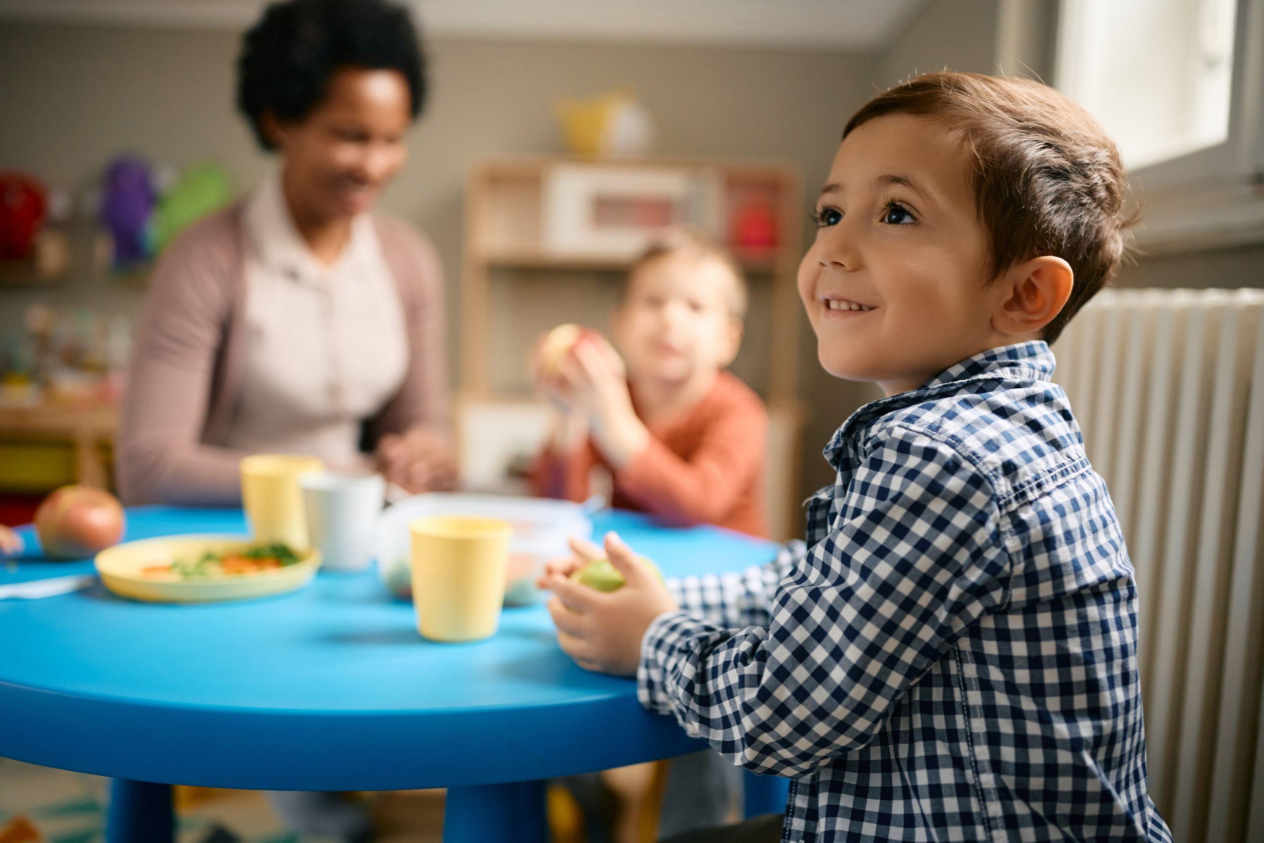 A smiling child in a checkered shirt sits at a blue table with food.