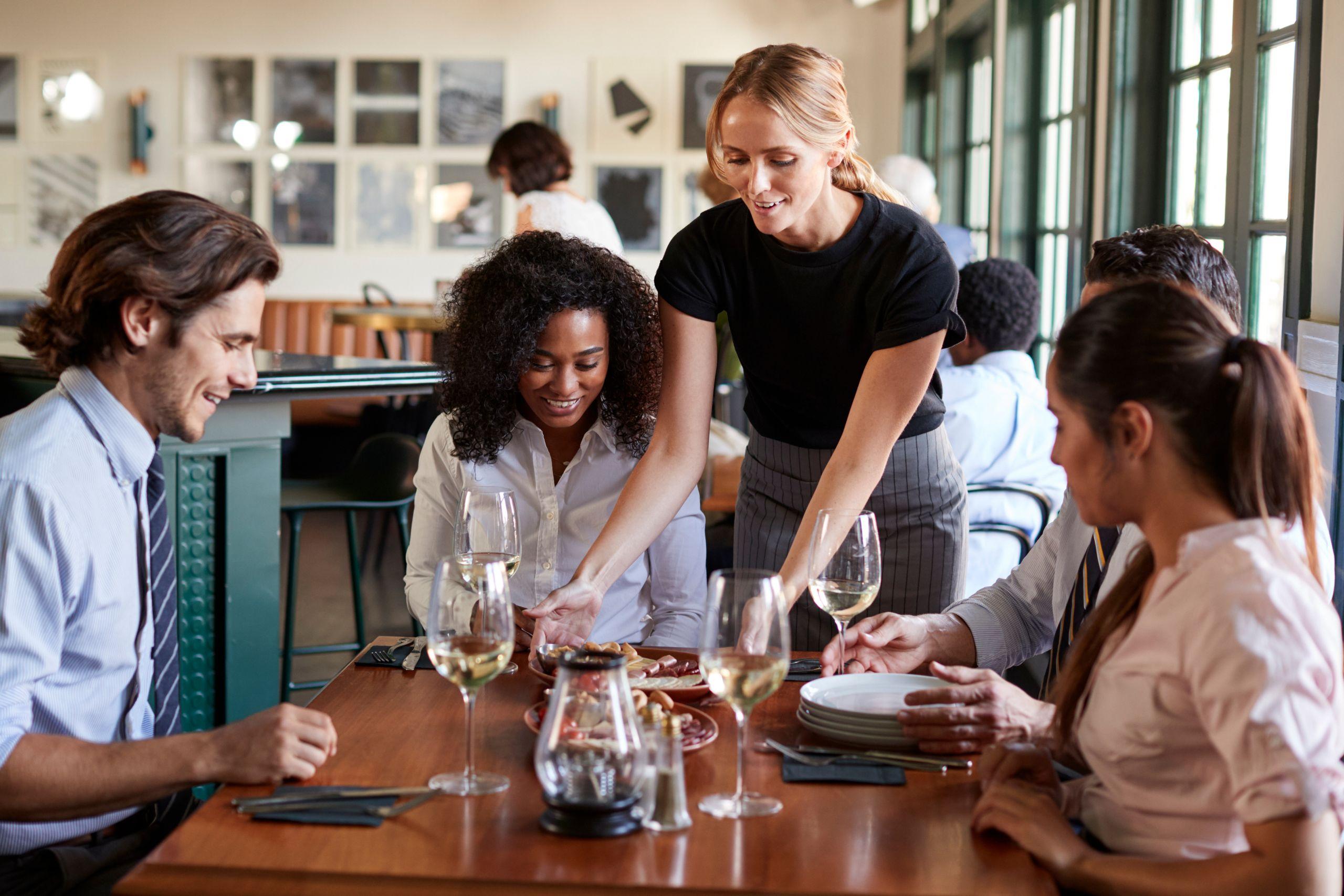 A waitress serves food to a group of four people sitting at a restaurant table with wine glasses, engaging in conversation.