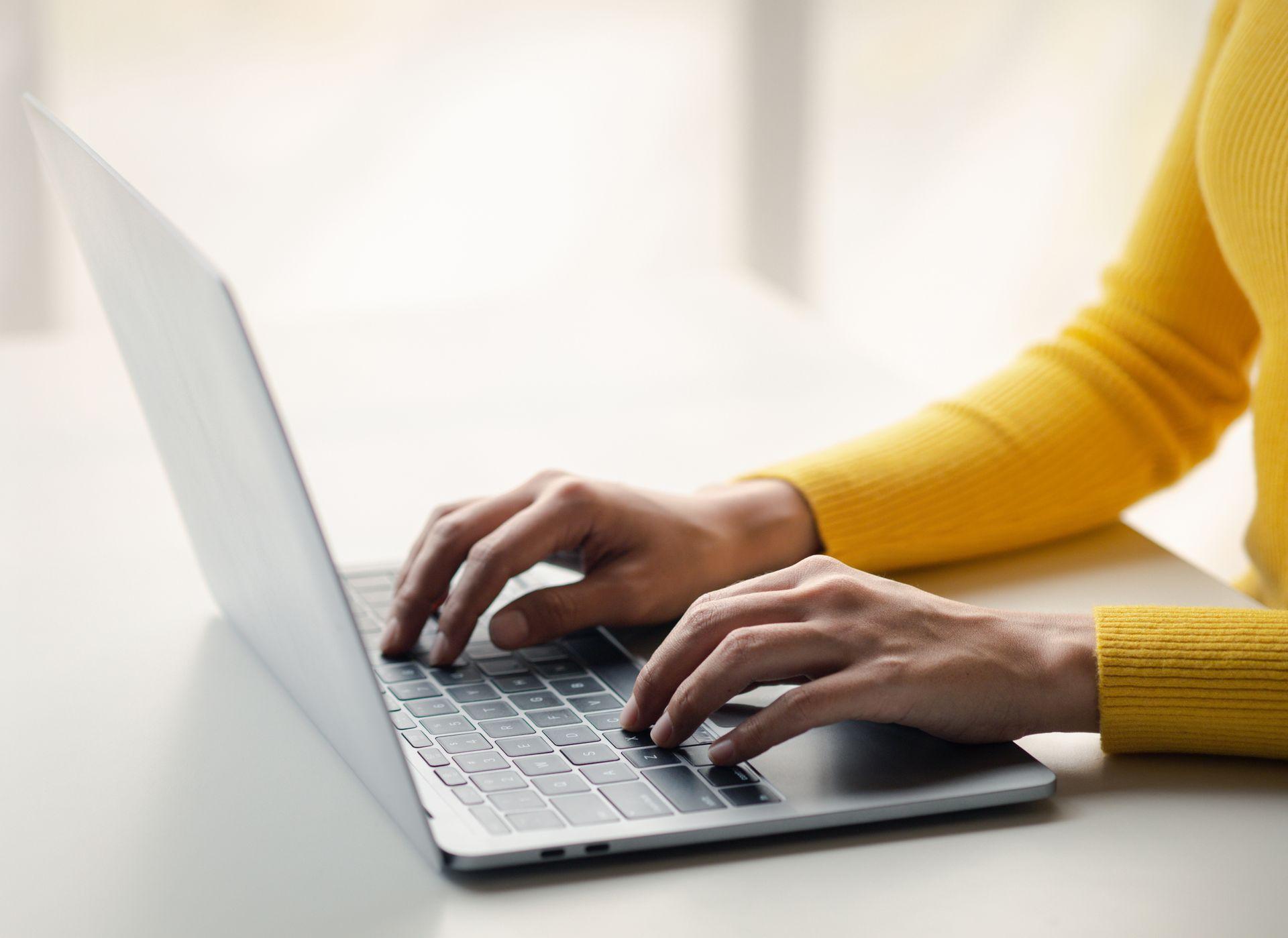 Person in a yellow sweater typing on a laptop keyboard on a white table.