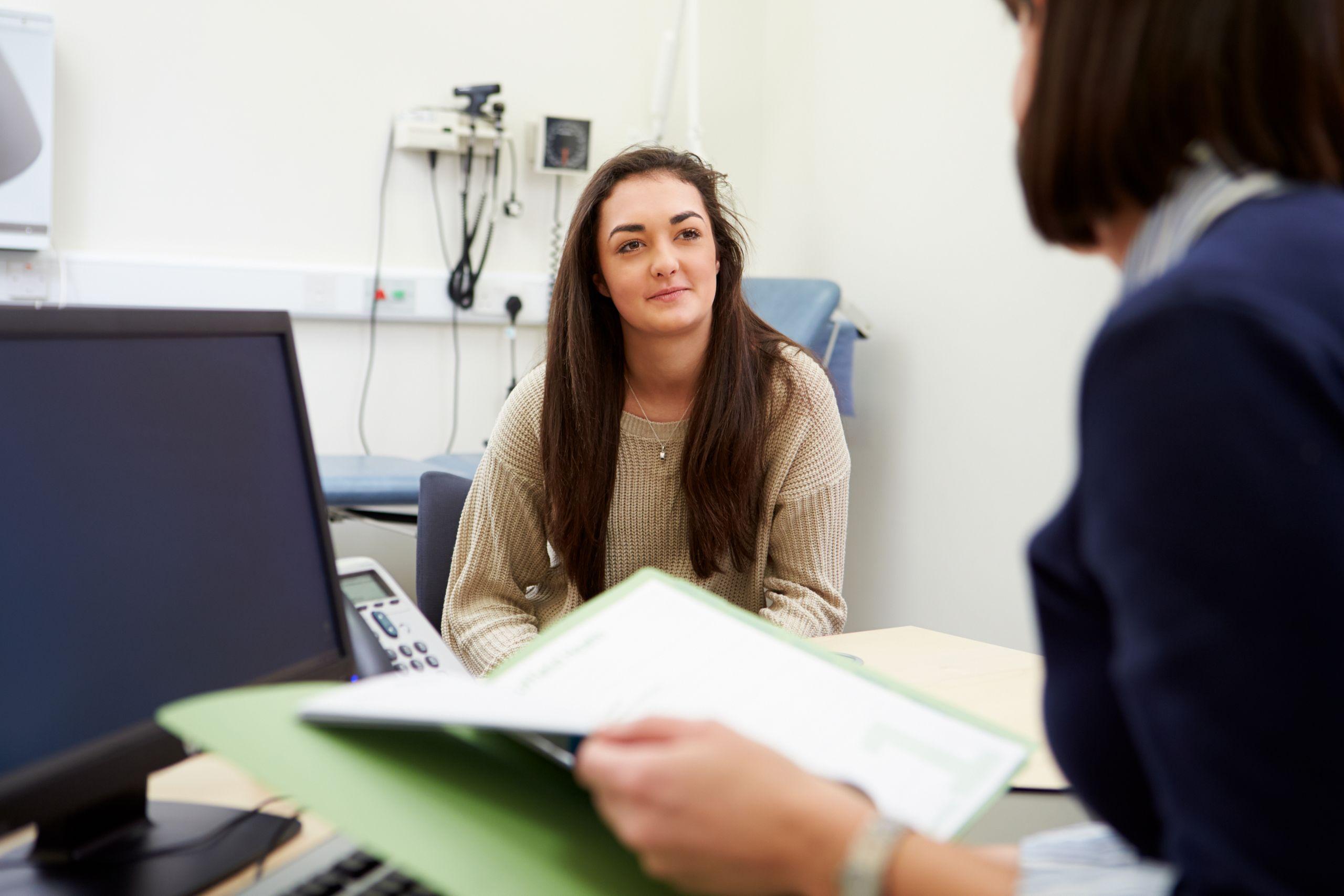 A woman sits across from a person holding a folder in an office setting, possibly discussing something. A computer and medical equipment are in the background.
