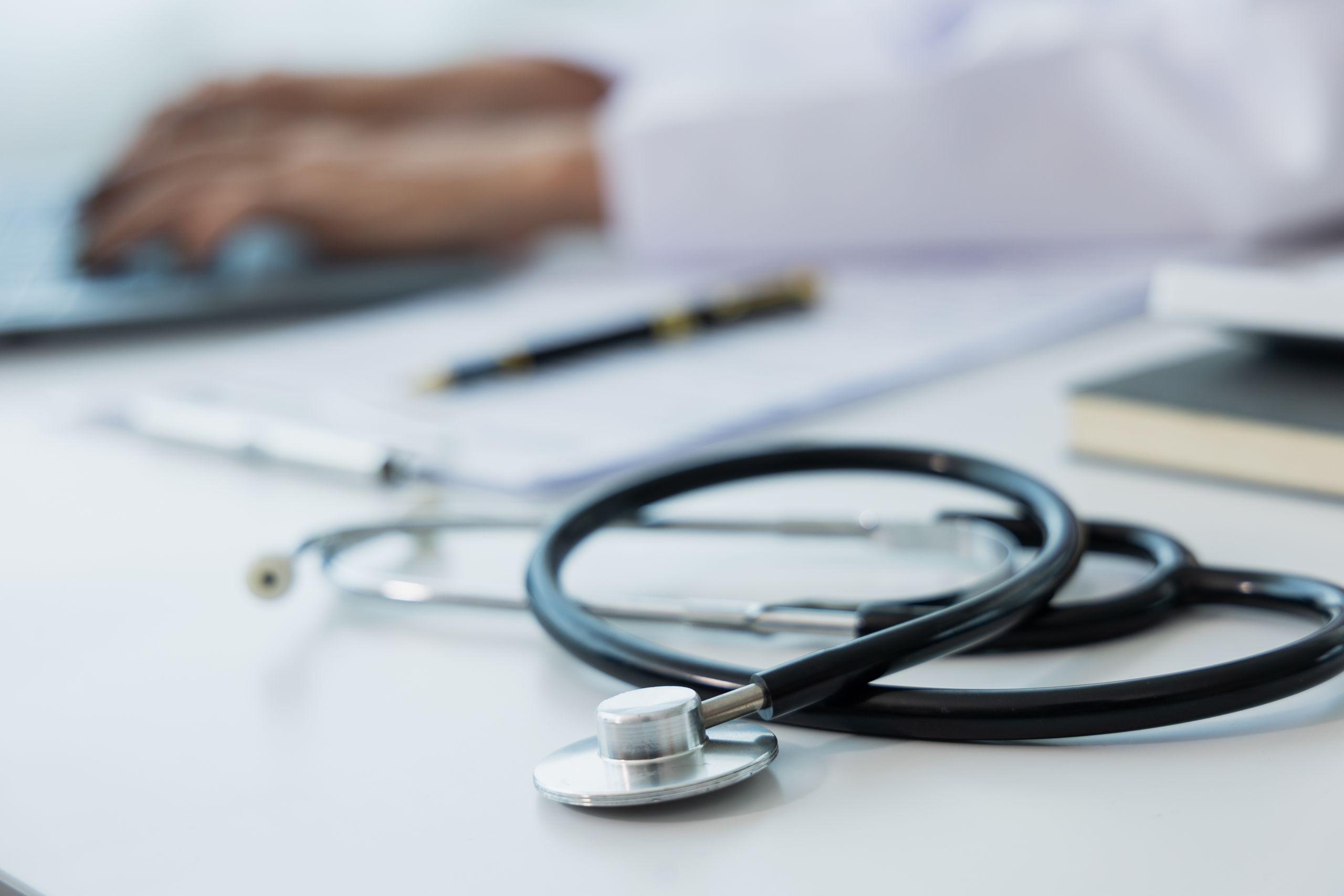 Stethoscope on a desk with a laptop keyboard in use by blurred hands, a pen and notebook in the background.