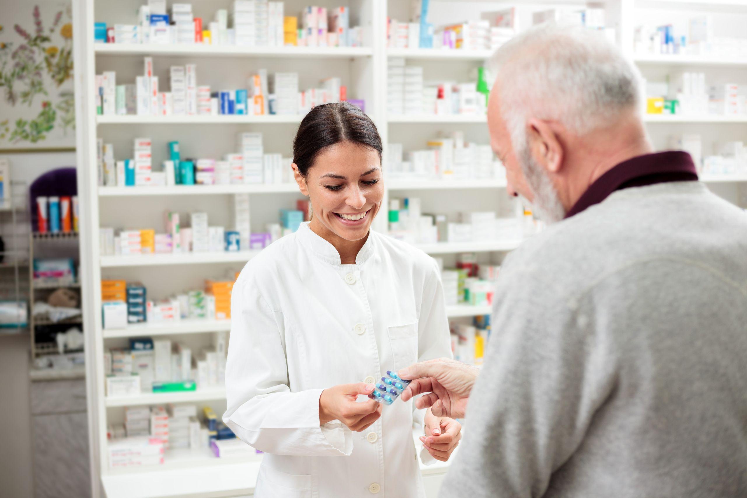 A pharmacist in a white coat hands medication to an elderly customer in a pharmacy with shelves of medicine in the background.