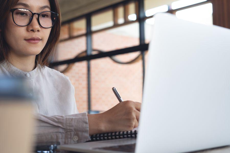 Person wearing glasses, writing in a notebook while looking at a laptop screen, with a blurred background of windows and a brick wall.