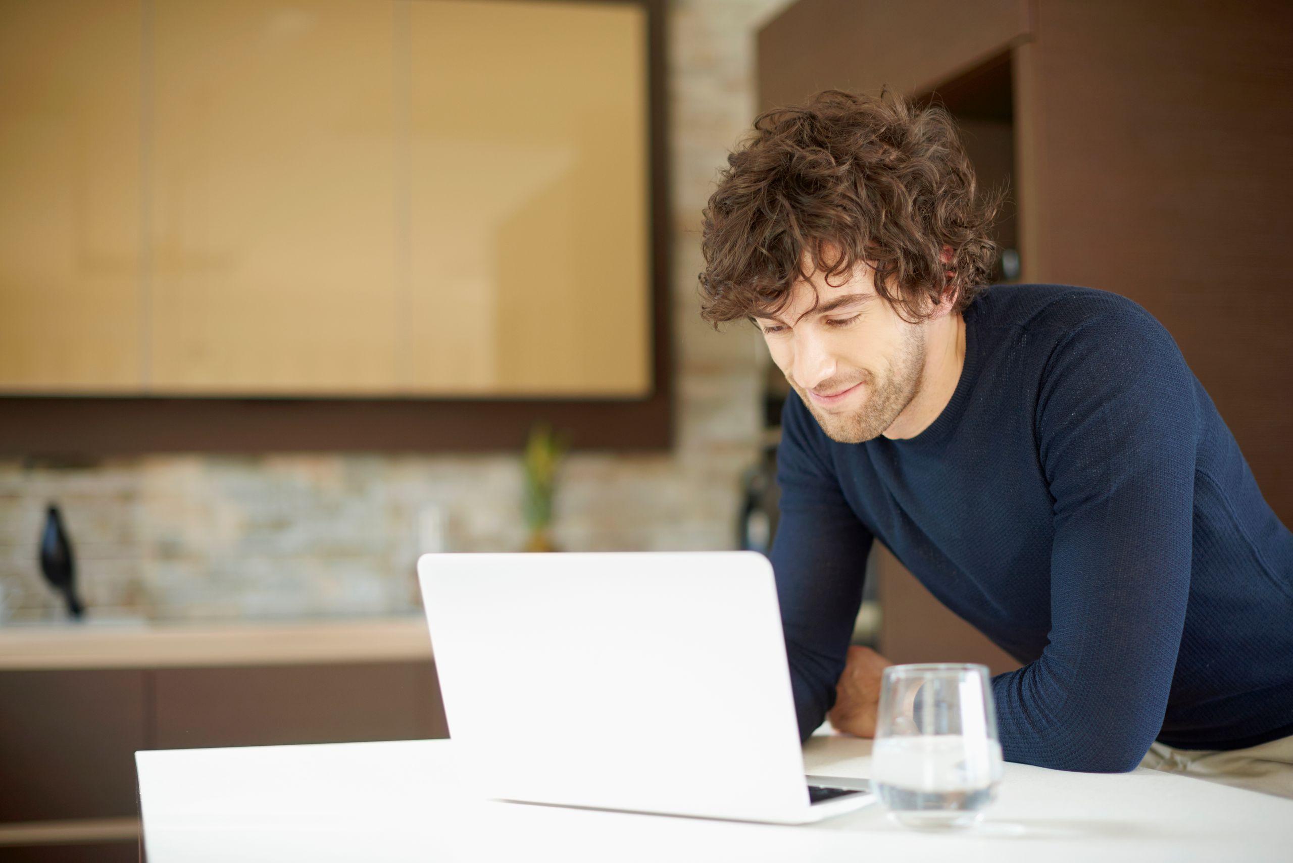 Man with curly hair leaning over a laptop on a table, smiling, in a modern kitchen setting. A glass of water is on the table.