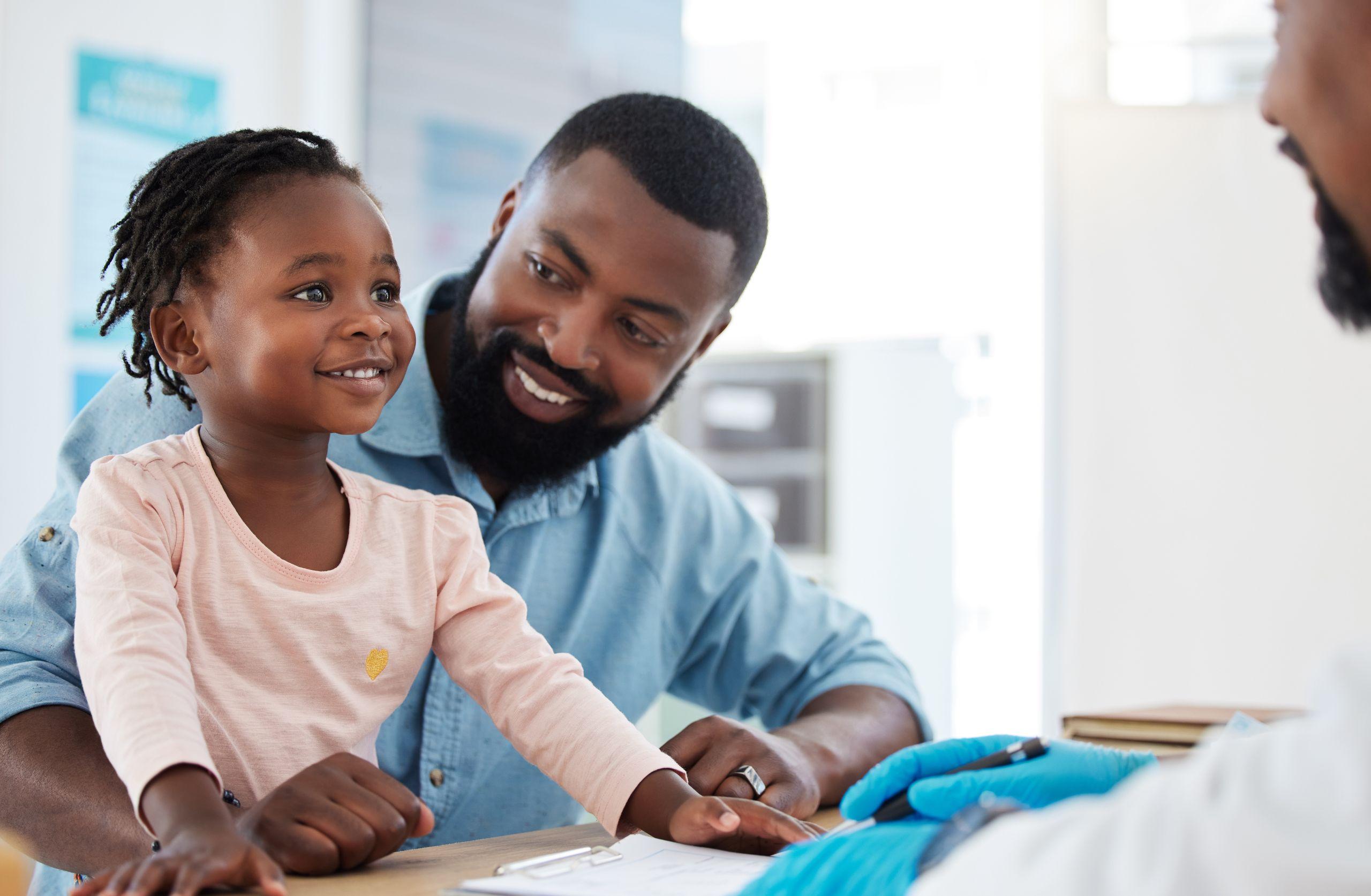 A father and his smiling child engage with a healthcare professional in an office setting. The child is sitting on a table, appearing happy.
